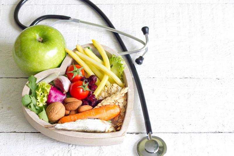 Heart-shaped bowl with vegetables, nuts, and fish alongside a stethoscope symbolizing healthy diet and medical nutrition therapy.