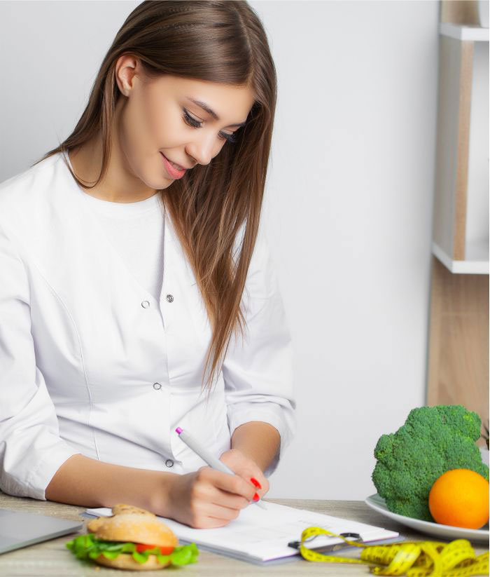 Female nutritionist writing personalized diet plan with healthy foods like broccoli, orange, and sandwich on the table.